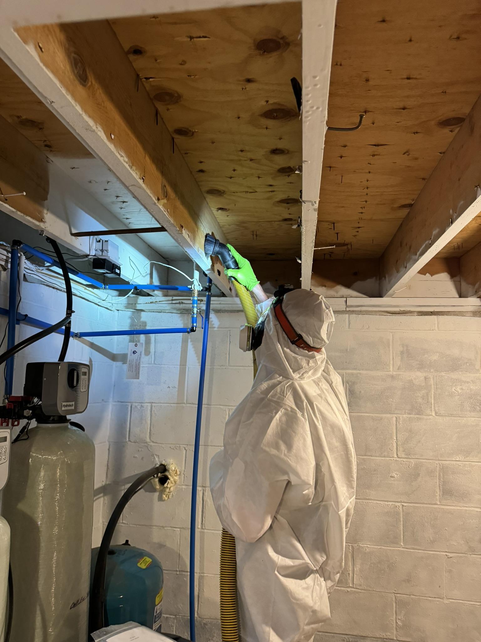 A certified technician in a hazmat suit using a HEPA vacuum on basement ceiling joists.